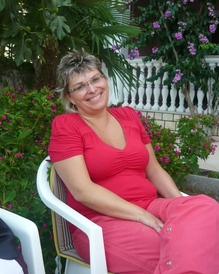 Femme assise sur une chaise, en robe rouge, souriant, entourée de fleurs.