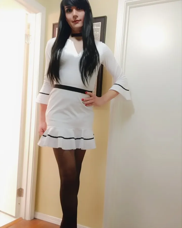 Femme aux cheveux noirs portant une robe blanche courte, bas noirs et talons aiguilles, posant dans un couloir.