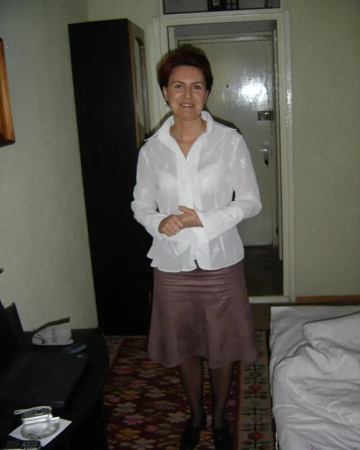 Femme aux cheveux châtains courts, en chemise blanche transparente et jupe marron, debout dans une chambre.
