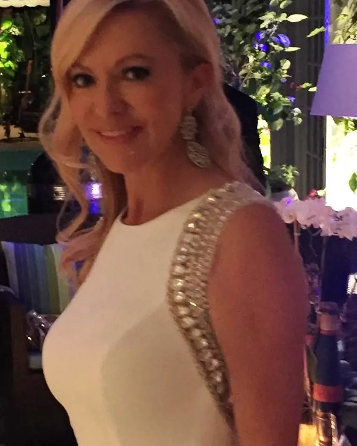 Femme blonde aux cheveux courts, en robe blanche, souriante, dans un cadre intérieur élégant.