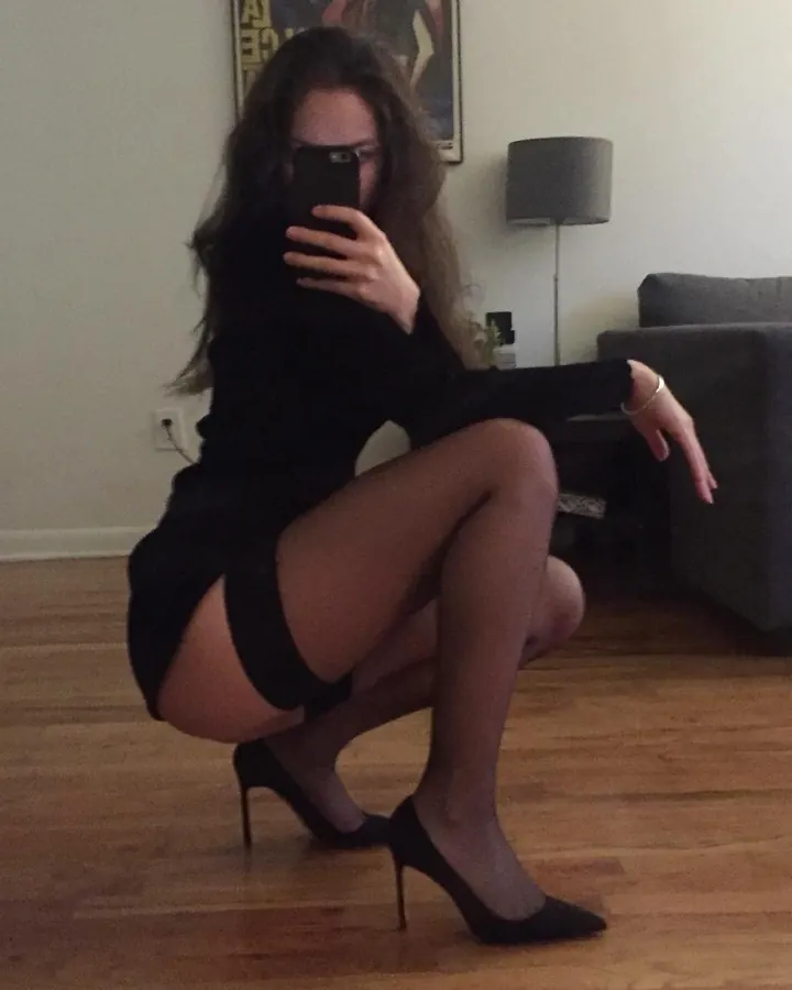 Femme aux cheveux bruns, en tenue noire, prenant un selfie en position accroupie dans un salon.