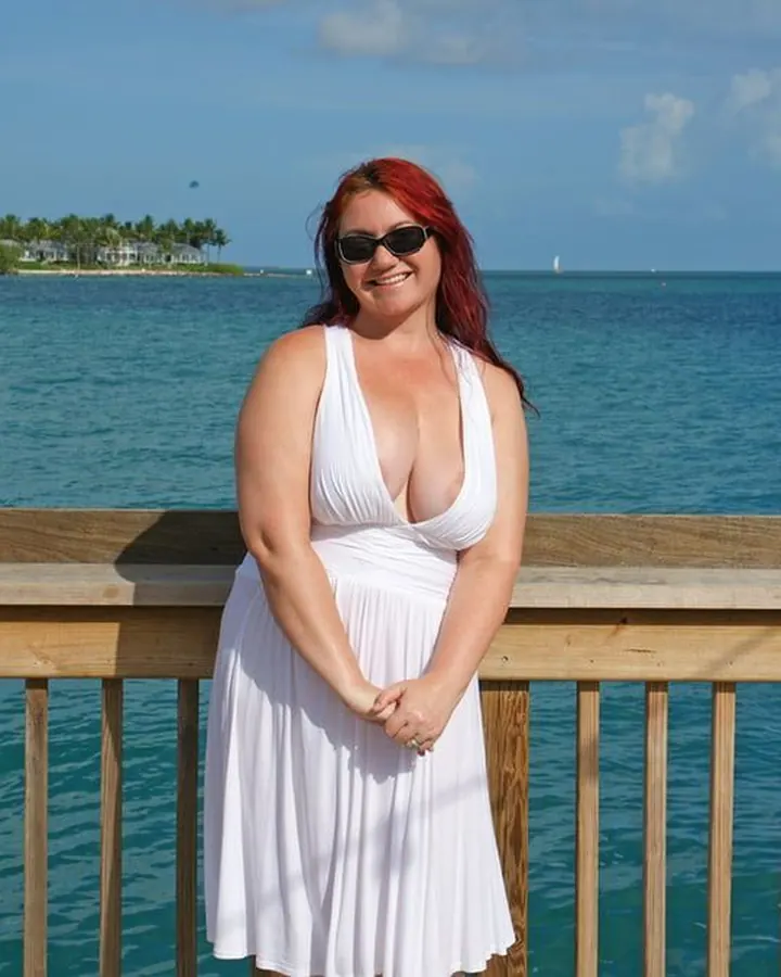 Femme aux cheveux roux, en robe blanche, souriante, devant un lac sous un ciel bleu.