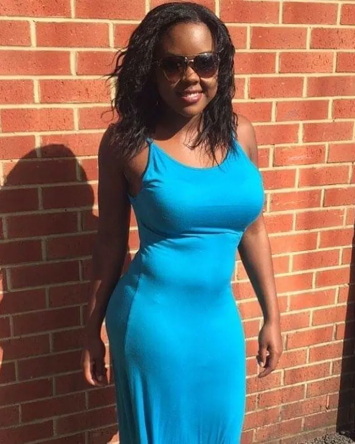 Femme noire aux cheveux bruns, en robe bleue, souriante, appuyée contre un mur en briques.