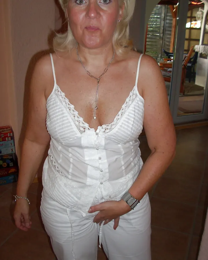 Femme blonde debout, en haut blanc à dentelle et pantalon blanc, regardant l'appareil
