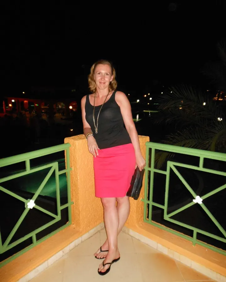 Femme blonde en tenue de soirée, debout sur un balcon la nuit, avec une jupe rose et un haut noir