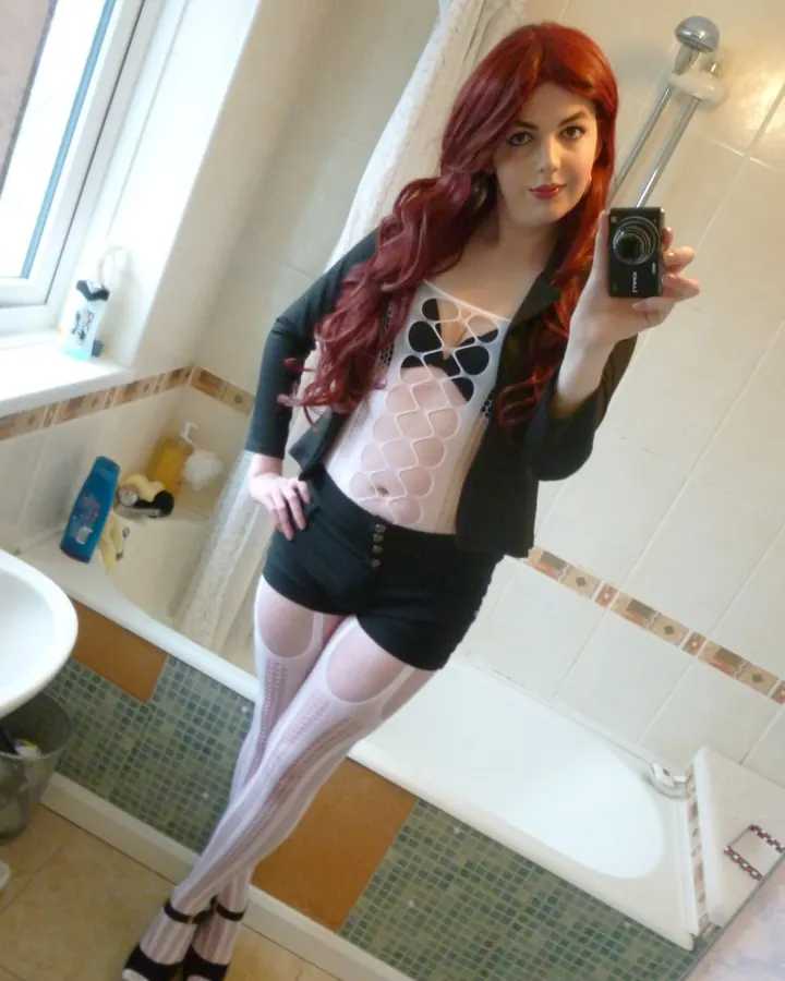 Femme aux cheveux roux en haut noir et jupe courte, prenant un selfie dans une salle de bain.