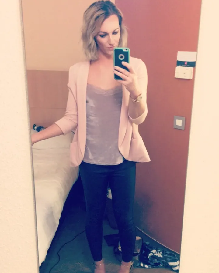 Femme blonde en veste rose pâle et leggings noirs, prenant un selfie dans une chambre.