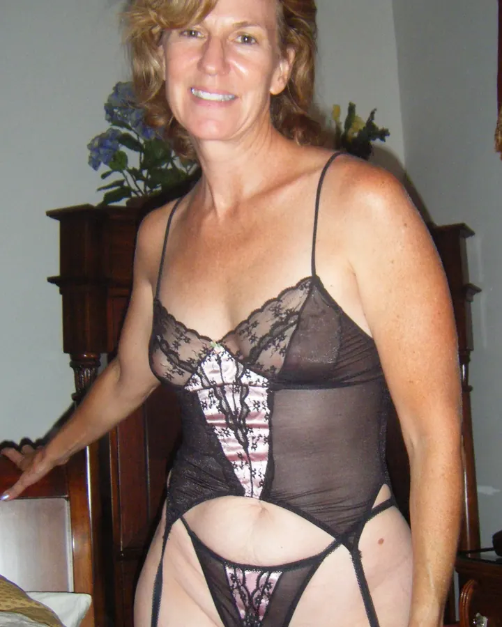Femme en lingerie noire transparente, debout près d'un lit en bois sombre.