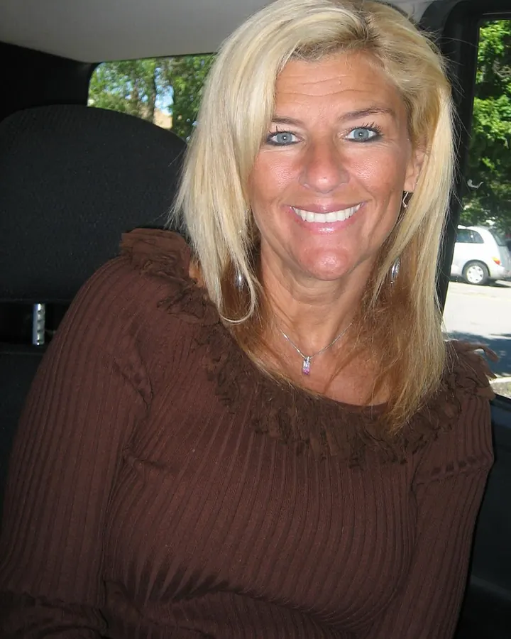 Femme blonde souriante en pull marron, assise à l'arrière d'une voiture.