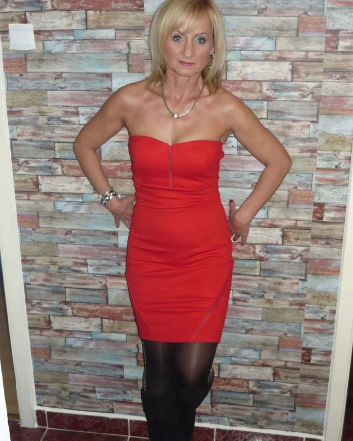 Femme blonde en robe rouge moulante, pose les mains sur les hanches devant un mur en bois.