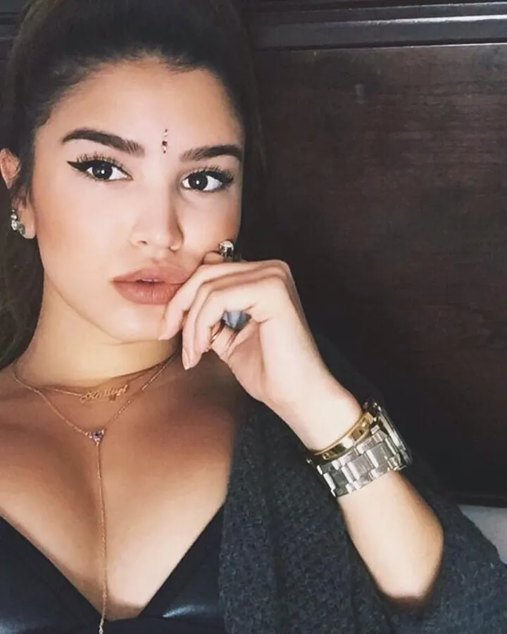 Femme latine aux cheveux bruns-noirs, maquillée, avec un point rouge entre les sourcils, portant un haut noir et une cardigan grise, assise sur un lit, main près de la bouche.