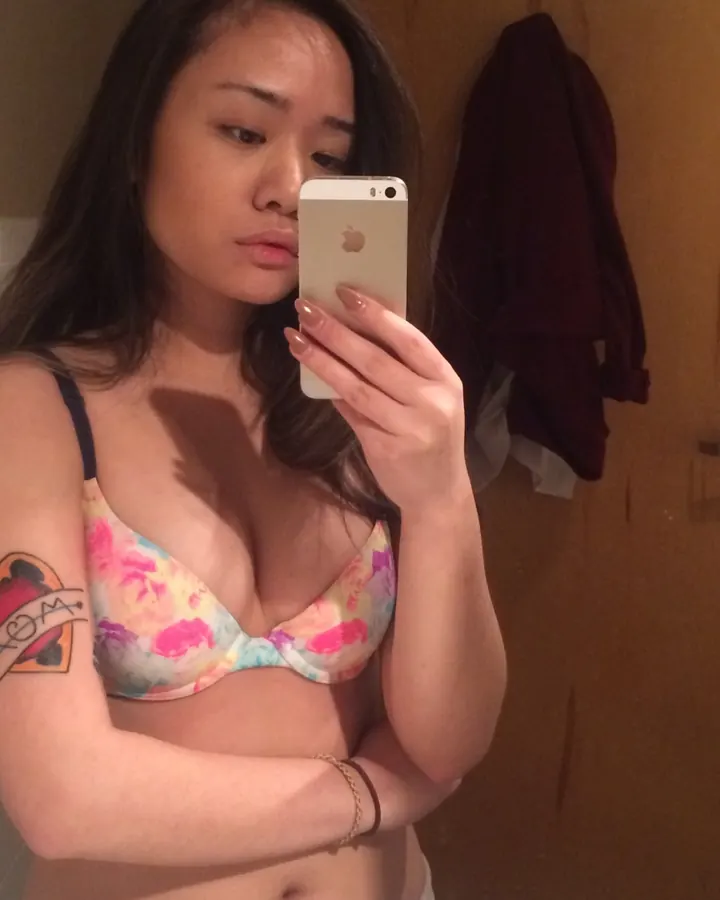 Jeune femme asiatique aux cheveux bruns-noirs, regard pensif, tenant un téléphone blanc, portant un soutien-gorge fleuri coloré, dans un cadre intérieur avec porte en bois.