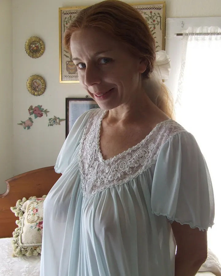 Femme européenne aux cheveux roux, souriante, portant une chemise de nuit bleu clair avec dentelle blanche, posant dans une chambre avec lit et décor floral.