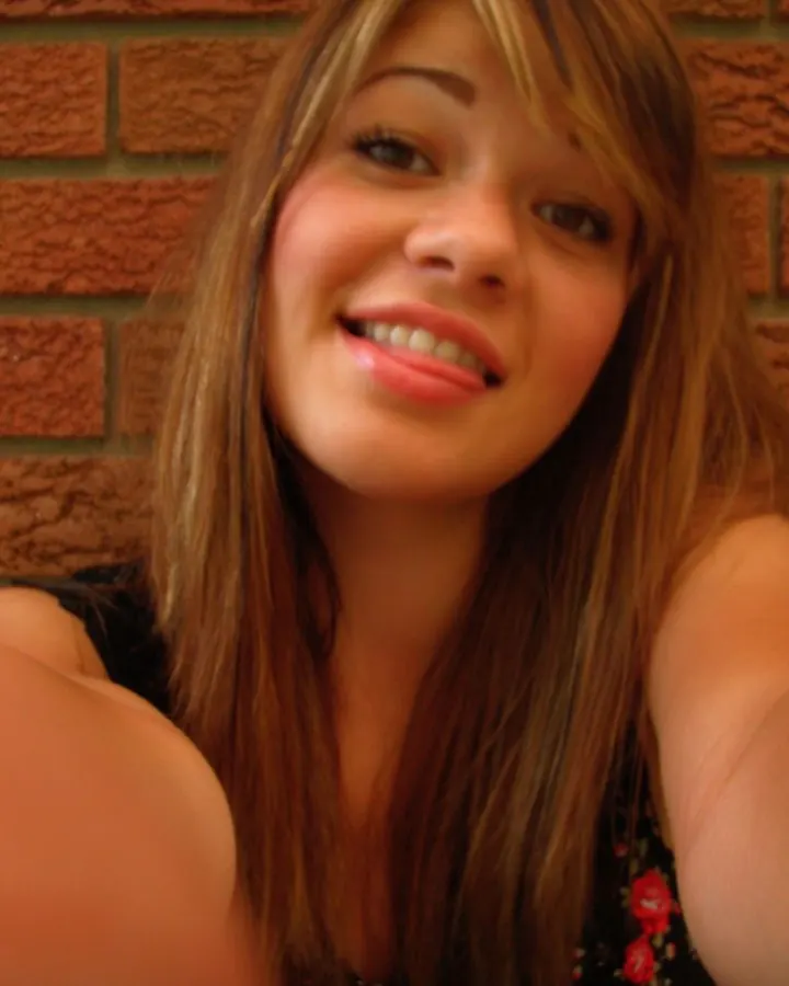 Jeune femme aux cheveux châtains, souriante, prenant un selfie avec un téléphone, portant un haut noir et un short en jean, dans une pièce avec un téléviseur en arrière-plan.