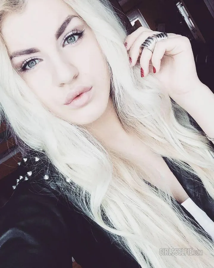 Jeune femme aux cheveux blonds et yeux bleus, regard intense, portant une veste noire ornée de clous, tenant sa chevelure blonde avec une main et un anneau à l'index.