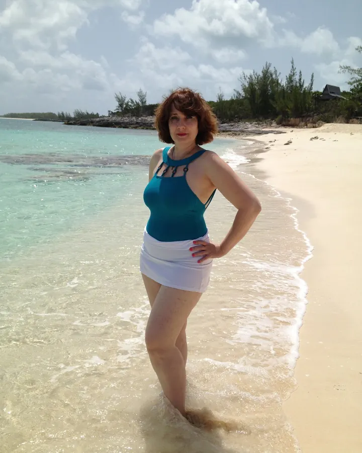 Femme en maillot turquoise et short blanc, debout dans l'eau claire sur une plage.