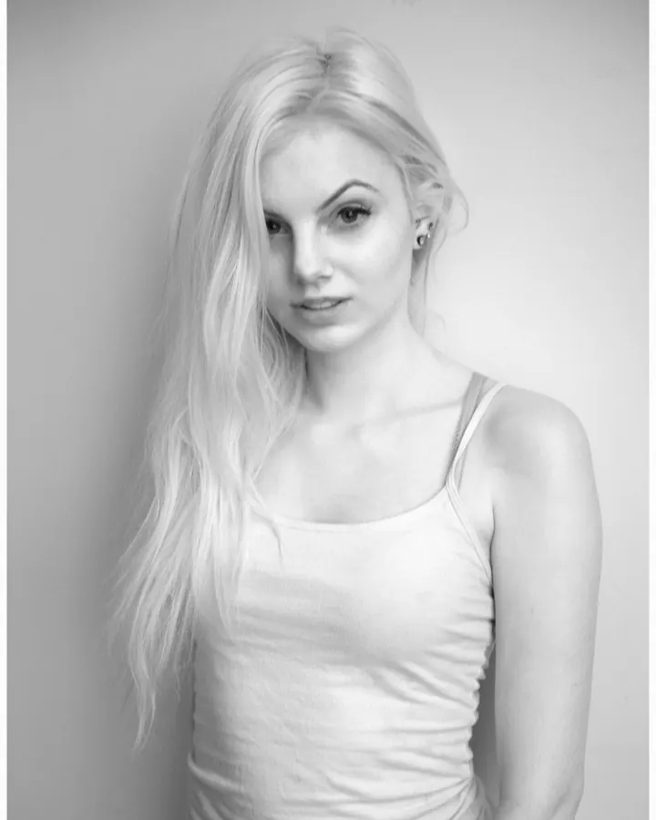 Jeune femme blonde aux cheveux mi-longs, souriante, portant un haut jaune et un short blanc, debout devant une cuisinière blanche, les mains derrière le dos.