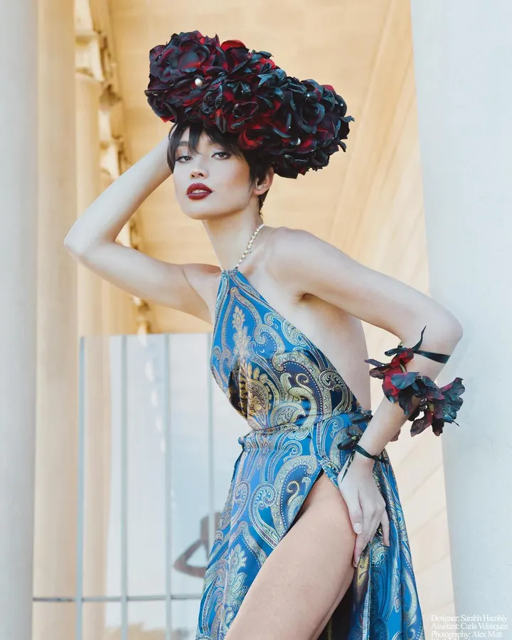 Jeune femme asiatique aux cheveux bruns-noirs, portant une robe bleue à motifs dorés, avec un grand chapeau de fleurs rouges et noires, posant dans un décor classique.