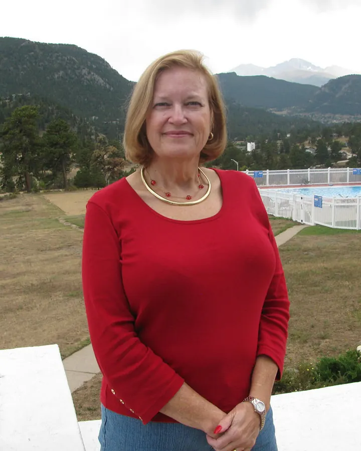 Femme aux cheveux blonds mi-longs, vêtue d’un haut rouge et d’un jean bleu, souriant doucement, debout sur un balcon avec une vue sur des montagnes et une piscine.