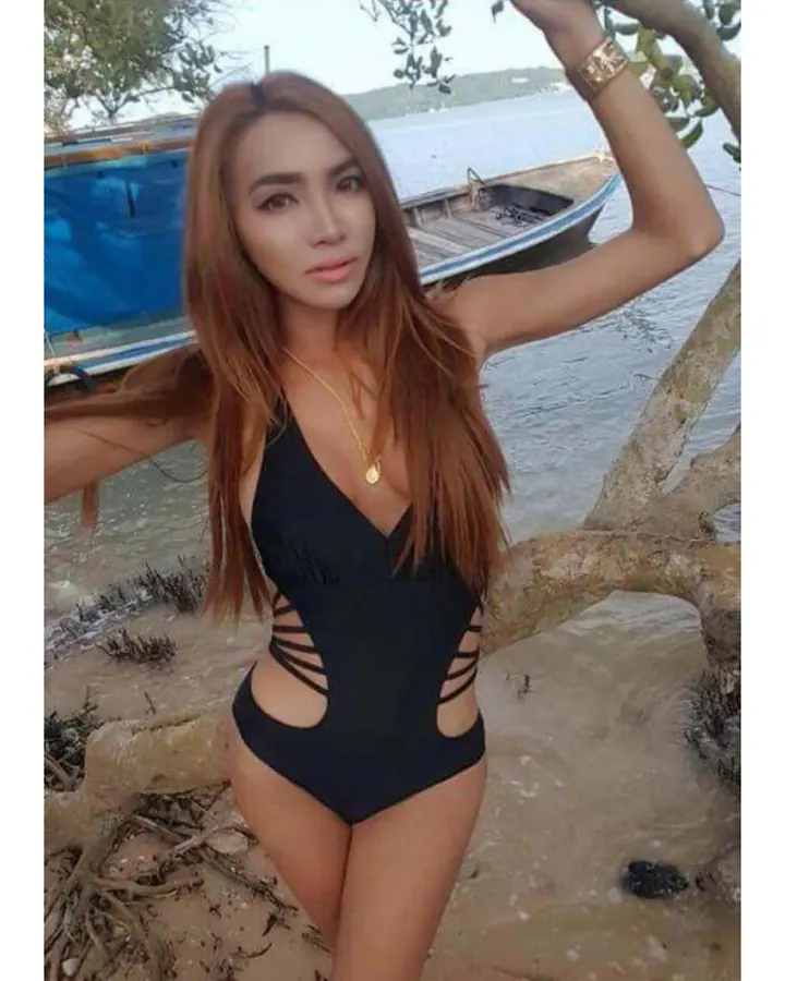 Femme aux cheveux châtains longs, vêtue d’un maillot de bain noir à découpes latérales, posant sur une plage avec un bateau en arrière-plan.