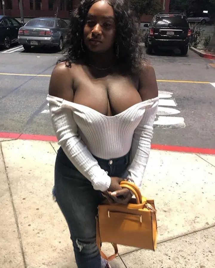 Femme aux cheveux bruns longs, vêtue d’un haut noir et d’un short blanc, posant dans un couloir avec un sourire large.