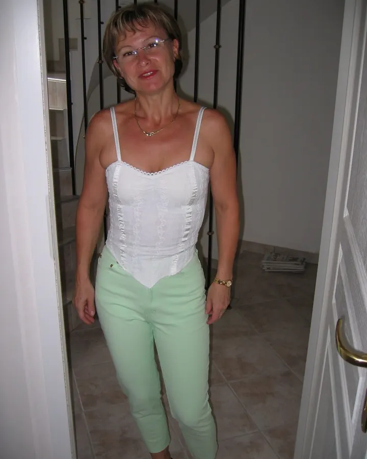 Femme aux cheveux châtains courts, lunettes, chemisier blanc et pantalon vert clair, souriante dans un couloir intérieur.