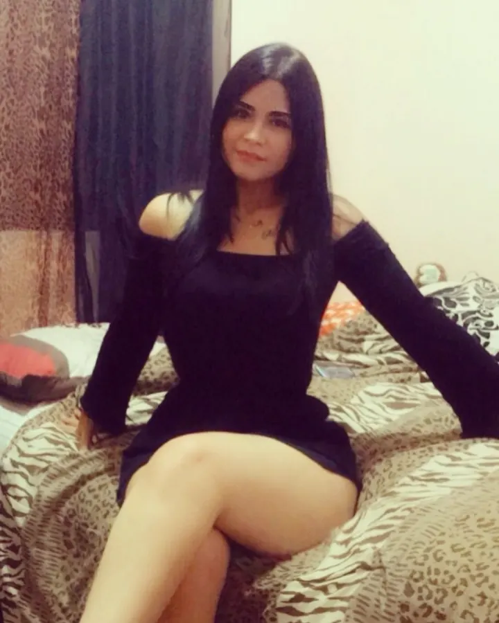 Femme aux cheveux noirs, en tenue simple, posant sur un lit, dans une chambre aux tons clairs.