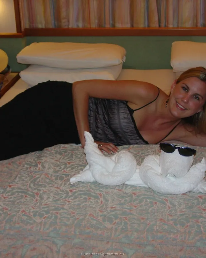 Femme aux cheveux blonds, allongée sur un lit en position latérale, souriant à l'appareil, avec deux serviettes pliées en forme d'animaux devant elle.