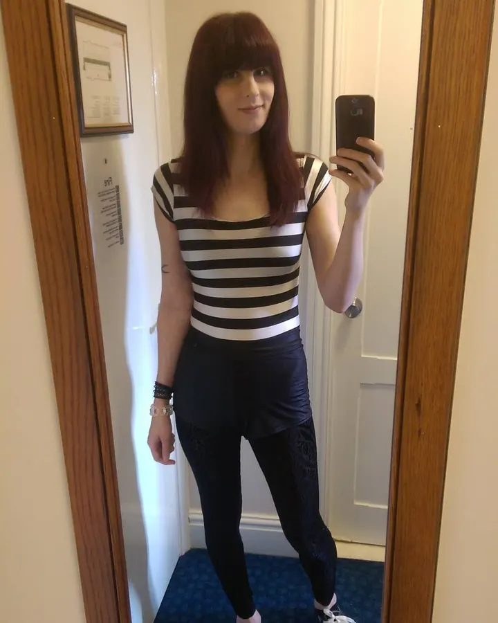 Femme aux cheveux châtains foncés et frange, en t-shirt rayé noir et blanc, prenant un selfie devant un miroir dans un couloir.