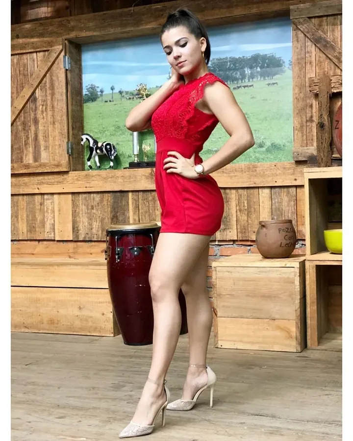 Femme aux cheveux bruns-noirs, en robe rouge ajustée, posant dans une pièce en bois avec un tambour en arrière-plan.