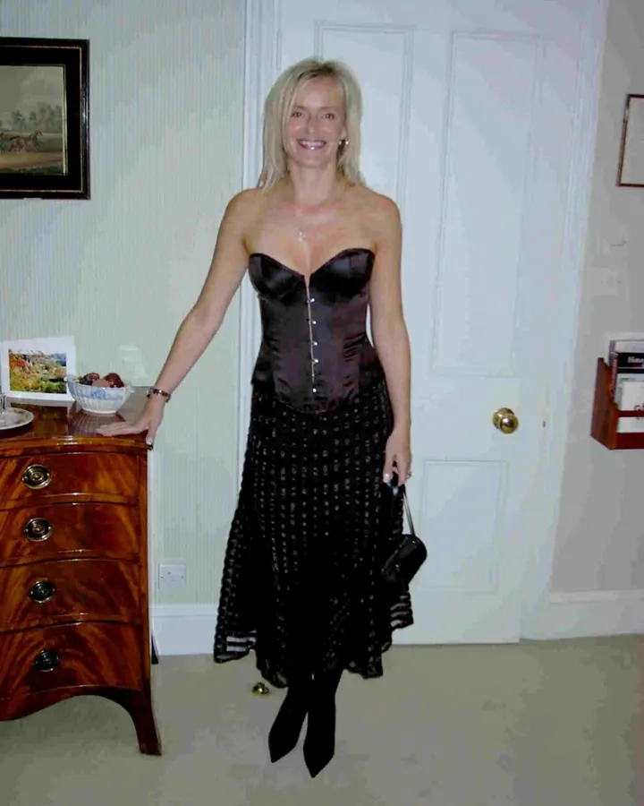 Femme aux cheveux blonds platine en robe noire, bustier, jupe longue, chaussures noires, tenant un sac, debout dans une pièce avec une commode en bois.