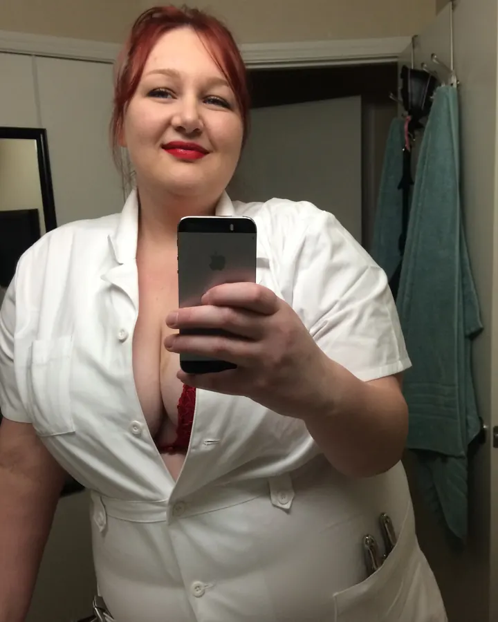 Femme rousse en blouse blanche médicale, soutien-gorge rouge visible, tenant téléphone, souriant
