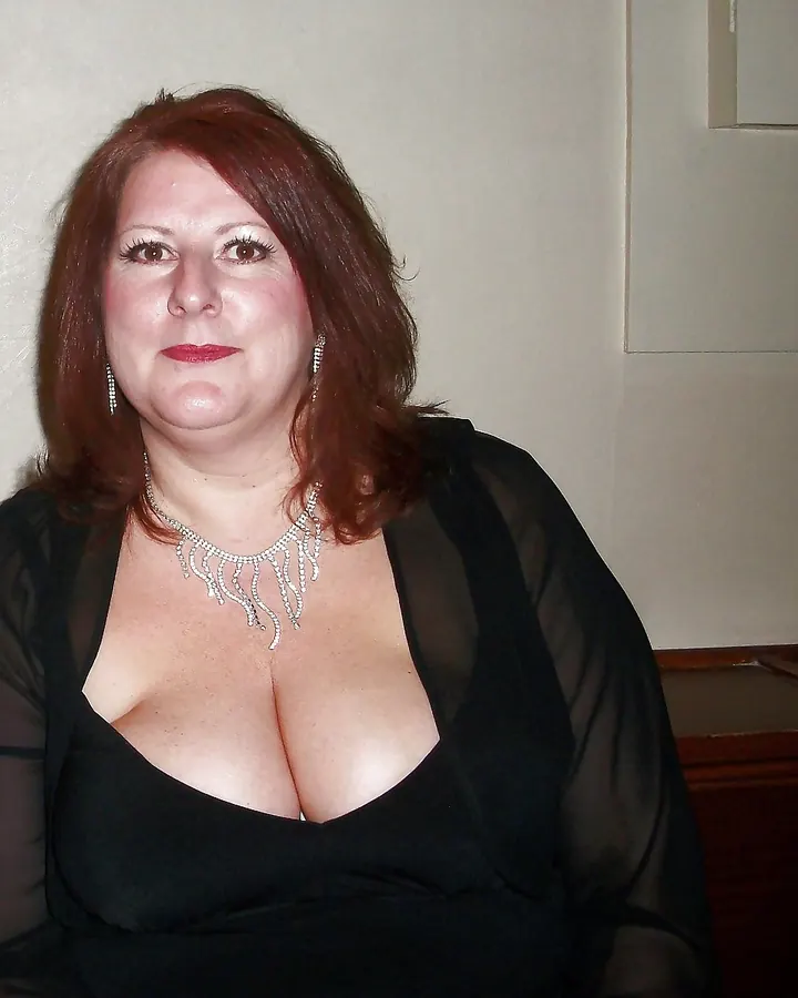 Femme rousse en robe noire transparente, collier pailleté, regard fixe, sourire discret
