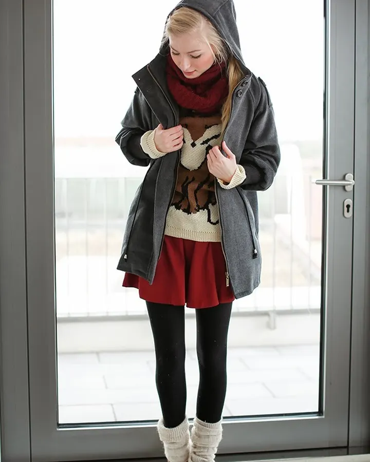 Jeune femme blonde portant un manteau gris, un pull beige à motif animalier, une jupe rouge, des collants noirs et des bottines marron, debout devant une porte vitrée.