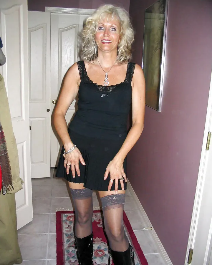 Femme aux cheveux platine portant une robe noire, posant en intérieur avec un sourire.