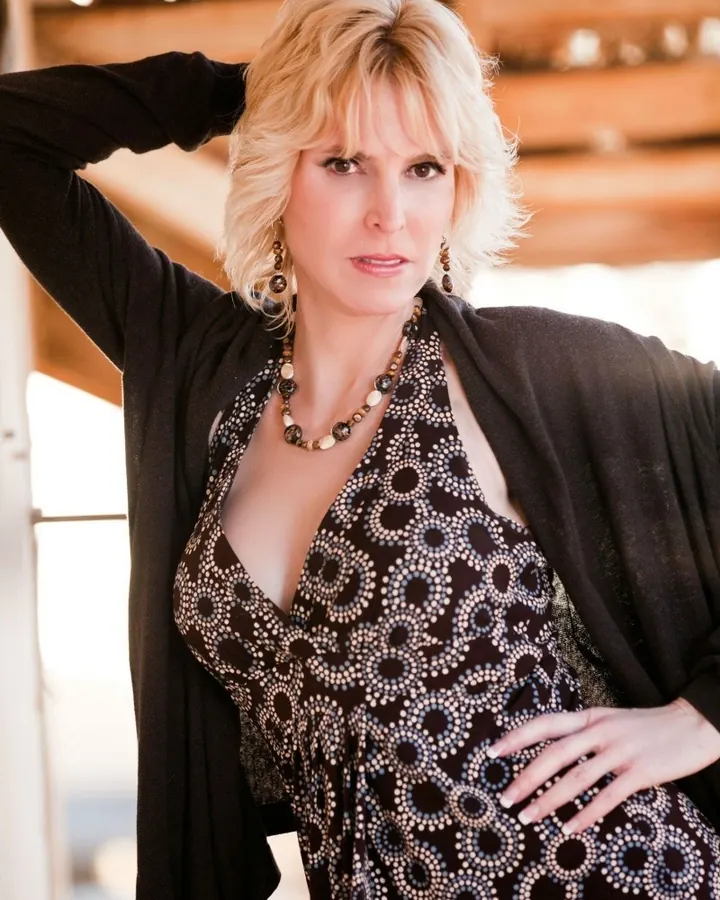 Femme blonde portant une robe noire à motifs circulaires et un cardigan noir, posant en extérieur sous une lumière chaude.