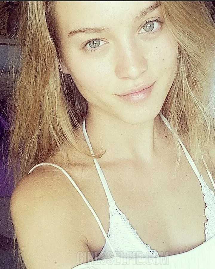 Femme blonde en soutien-gorge blanc, regardant l'appareil, cheveux soufflés par le vent, fond intérieur sombre.