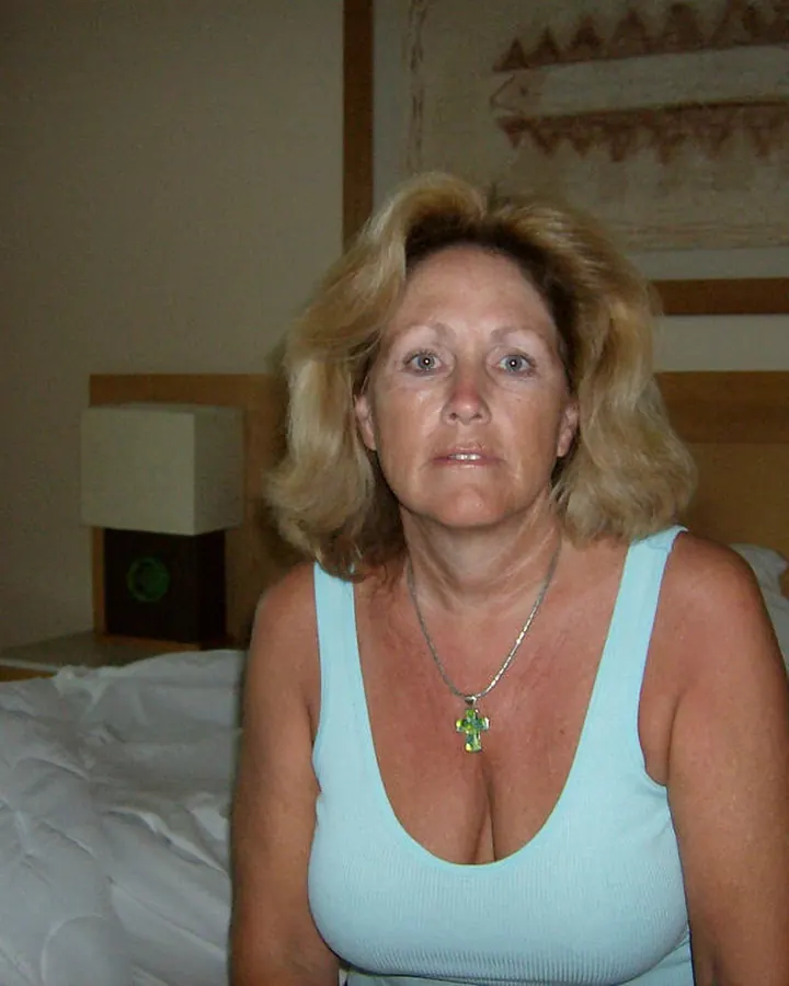 Femme aux cheveux blonds clairs portant un haut turquoise, assise sur un lit, regardant l'appareil.