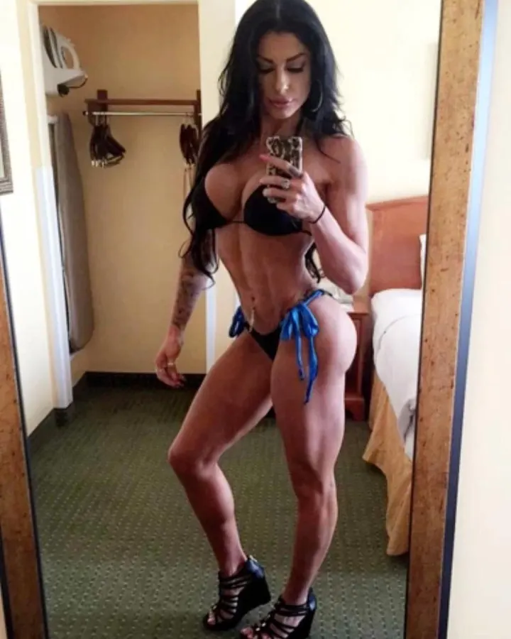 Femme aux cheveux noirs en bikini noir et blanc, posant contre un mur blanc, main sur la hanche, regardant l'appareil.