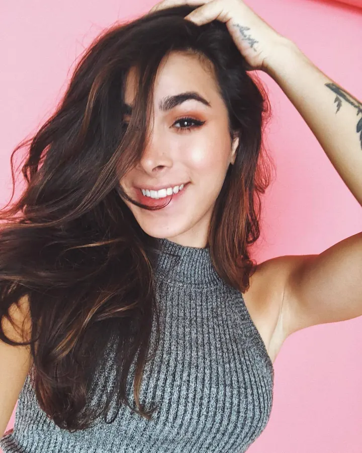 Femme aux cheveux foncés souriant, main dans les cheveux, fond rose, portant un haut gris tricoté.