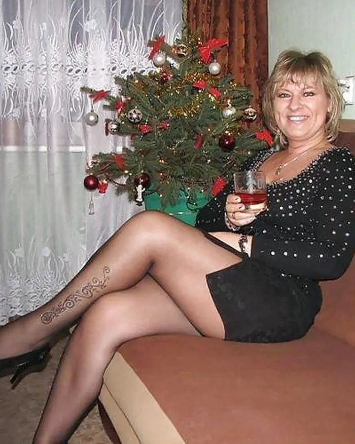 Femme blonde souriante, en robe noire, tenant un verre de vin rouge près d’un sapin de Noël