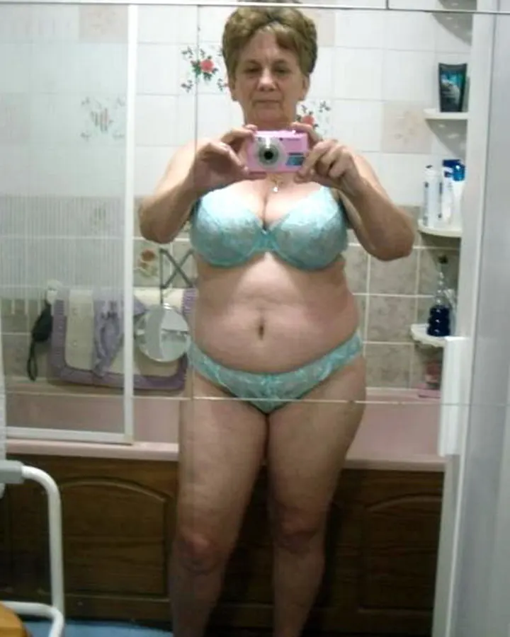 Femme âgée en sous-vêtements turquoise, prenant un selfie dans une salle de bain.
